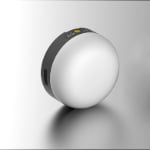 A round, white electronic device with a black band featuring buttons and a yellow circle, shown on a reflective surface.