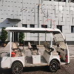 A white golf cart is parked on the street in front of a gray tiled building. A security camera, circled in red, is mounted high on the wall above the cart.