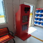 A red self-service kiosk with shelves stands indoors near a window, beside a bench and a rack of blue safety helmets.