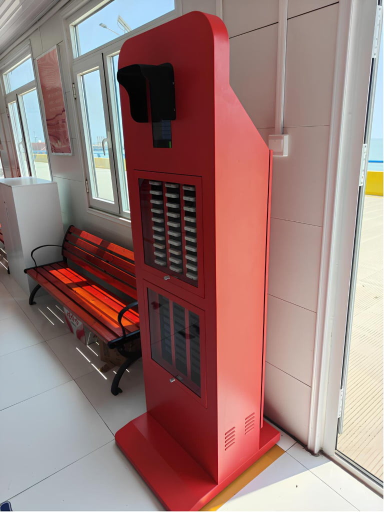 A tall red vending machine with shelves and a screen, placed indoors next to a window and an orange bench.