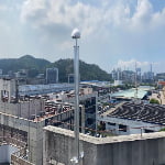 A rooftop with a tall metal pole topped by a dome-shaped device overlooks industrial buildings, with hills and a cityscape in the background under a partly cloudy sky.