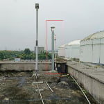 A vertical pipe with a capped top and attached wiring is positioned on a concrete rooftop near industrial storage tanks under an overcast sky.