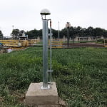 A metal pole with a dome-shaped device on top is mounted on a concrete base in a grassy outdoor area with industrial structures in the background.