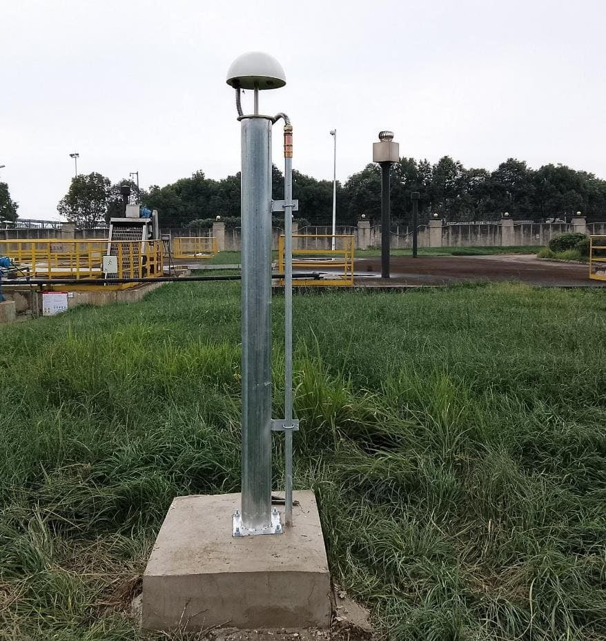 A metal pole with a dome-shaped device on top is mounted on a concrete base in a grassy outdoor area with industrial structures in the background.