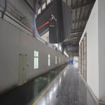 Interior of a large industrial facility with a long hallway, overhead structures, and an SN2 UWB ANCHOR alongside a security camera mounted high on the left wall highlighted by a red circle.