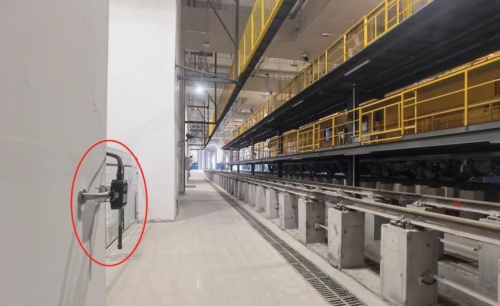 A maintenance bay with train tracks and yellow railings; a wall-mounted device inside a red circle is attached to pipes on the left.