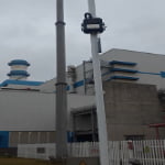 A white utility pole with a mounted black device stands in front of an industrial building with blue accents under a cloudy sky.