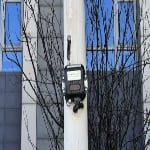 A black electronic device with an antenna is mounted on a white pole in front of a building with glass windows and leafless tree branches.