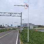A streetlight pole with a mounted camera circled in red stands near a road intersection, with a building and cloudy sky in the background.