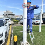 A worker in blue overalls stands on a ladder, installing a security camera on a white pole in a parking lot.