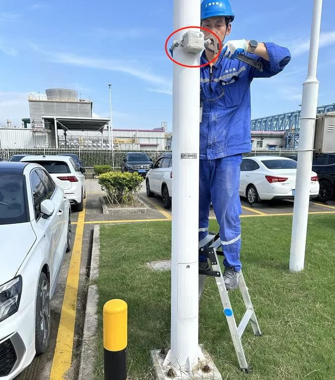 A worker in blue overalls stands on a ladder, installing a security camera on a white pole in a parking lot.
