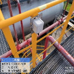 A gray electrical box is mounted on a red and yellow metal railing above an industrial area with large tanks. An information label with date, time, weather, and location is visible in the corner.