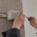 A person uses a wrench to adjust or install a small electrical box mounted on a textured wall.