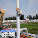 A sensor device is mounted on a white pole at an industrial facility, with pipes and buildings visible in the background. An information overlay displays date, time, weather, and location details.
