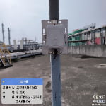 A metal device is mounted on a pole on an industrial rooftop. Information about the date, time, weather, and location is displayed in a blue box in the lower left corner.