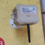 A rectangular wireless sensor device with a black antenna is mounted on a yellow wall. A label with Chinese text and numbers is visible on the front.