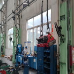 A red industrial machine part is suspended by straps from a crane inside a workshop. A worker wearing a blue helmet is at a workstation in the background.